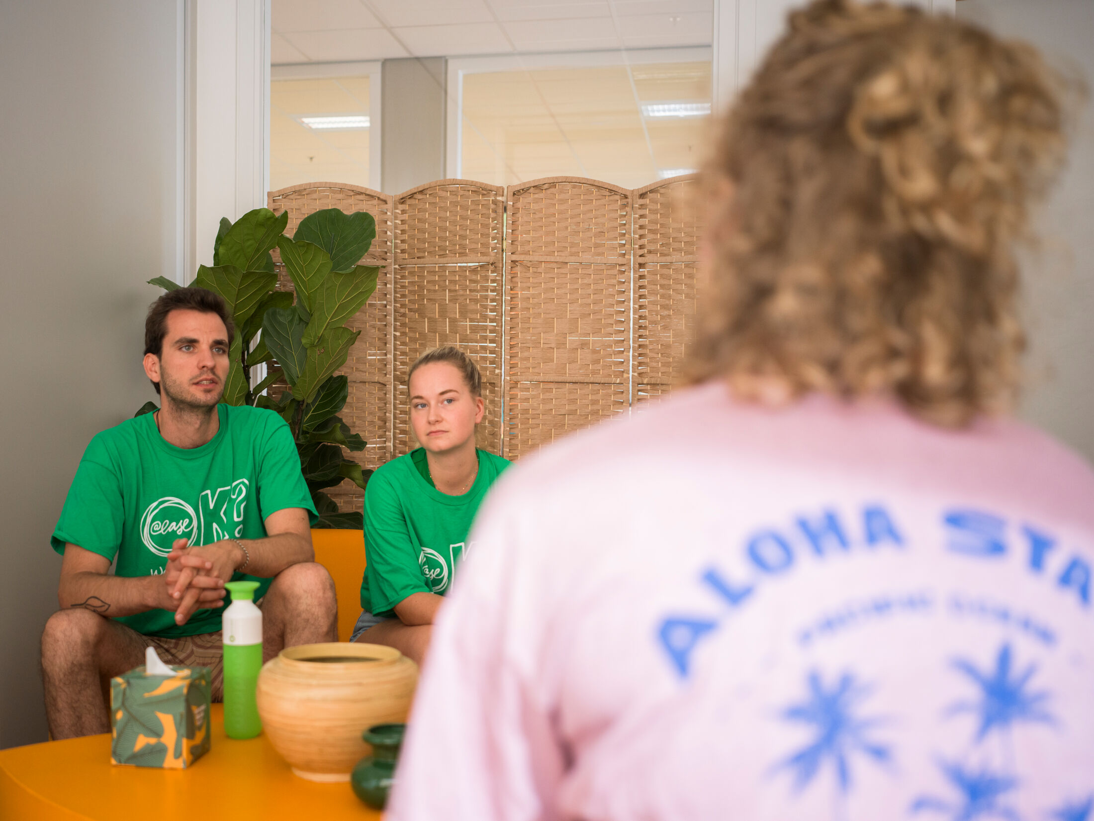 Twee vrijwilligers in een groen shirt van @ease luisteren naar iemand.