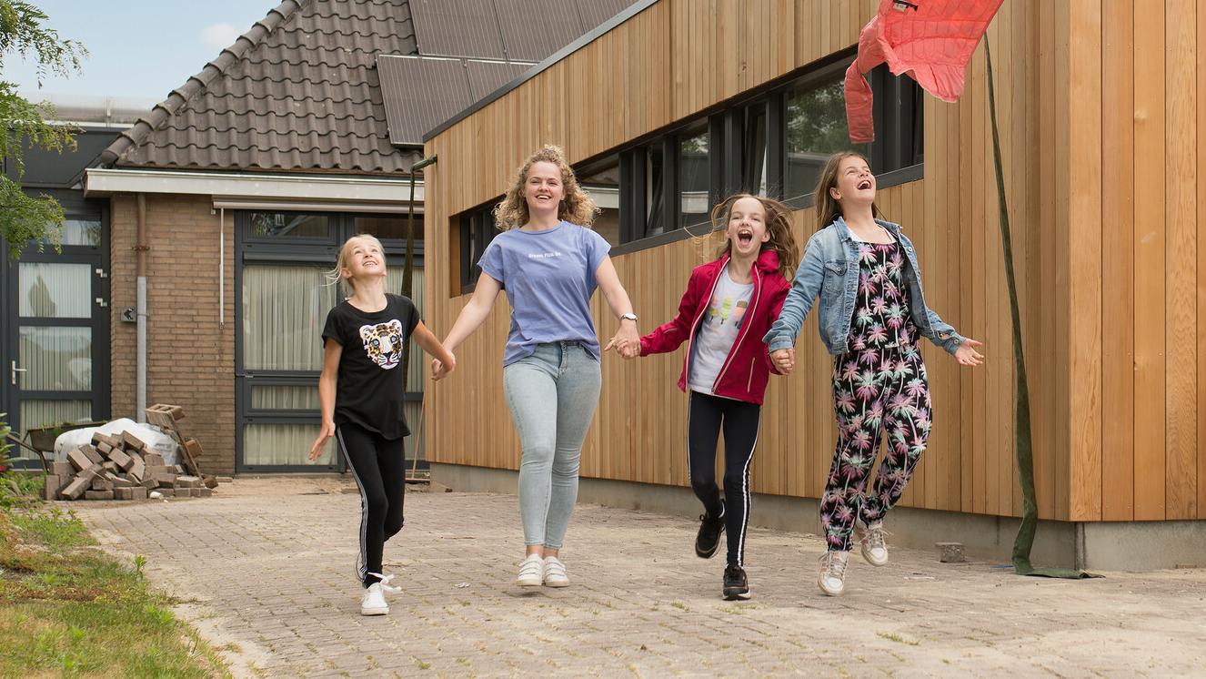 Een jongvolwassene loopt met drie kinderen blij een pad af. Ze houden elkaars hand vast.