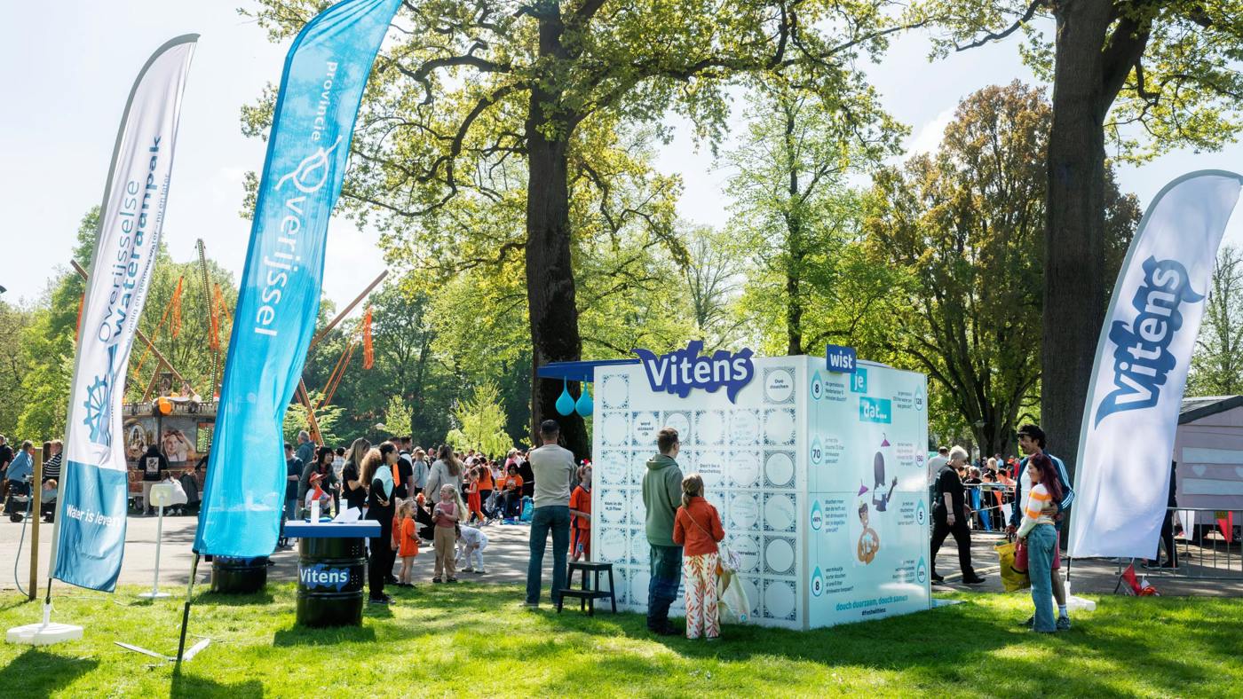 Mensen bezoeken een Vitens-stand met een douchekubus tijdens Koningsdag 2025 in het park, omringd door banners van Vitens en Provincie Overijssel.