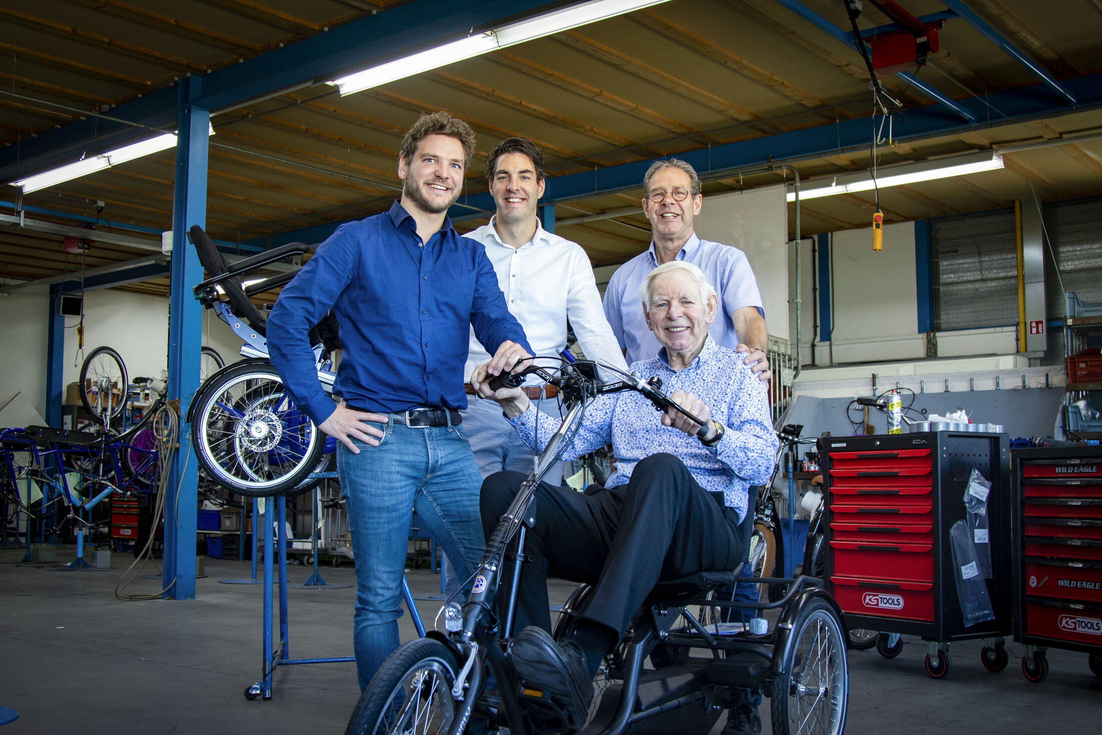 Drie mensen staan om een man die zit op een driewieler in een fietsenmakerswinkel