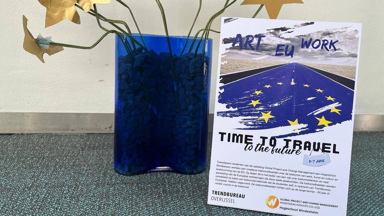 Een blauwe vaas met stenen en gouden bloemen staat naast een flyer met de titel "ART EU WORK TIME TO TRAVEL to the future". De flyer informeert over een evenement van studenten Global Project and Change Management aan Hogeschool Windesheim, over de toekomst van werk, kunst, cultuur en de EU.